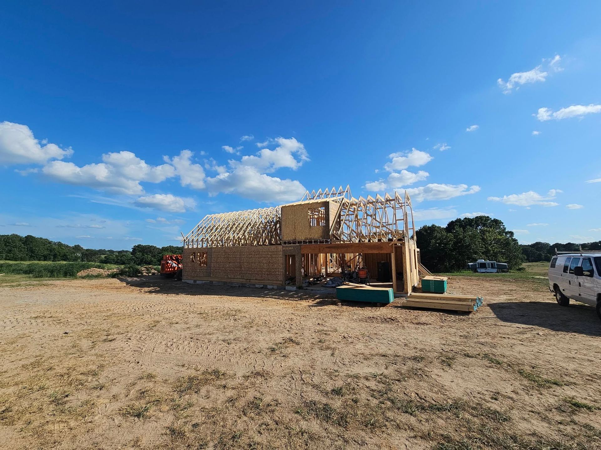 Partially framed wooden building on a dirt lot under a bright blue sky with clouds