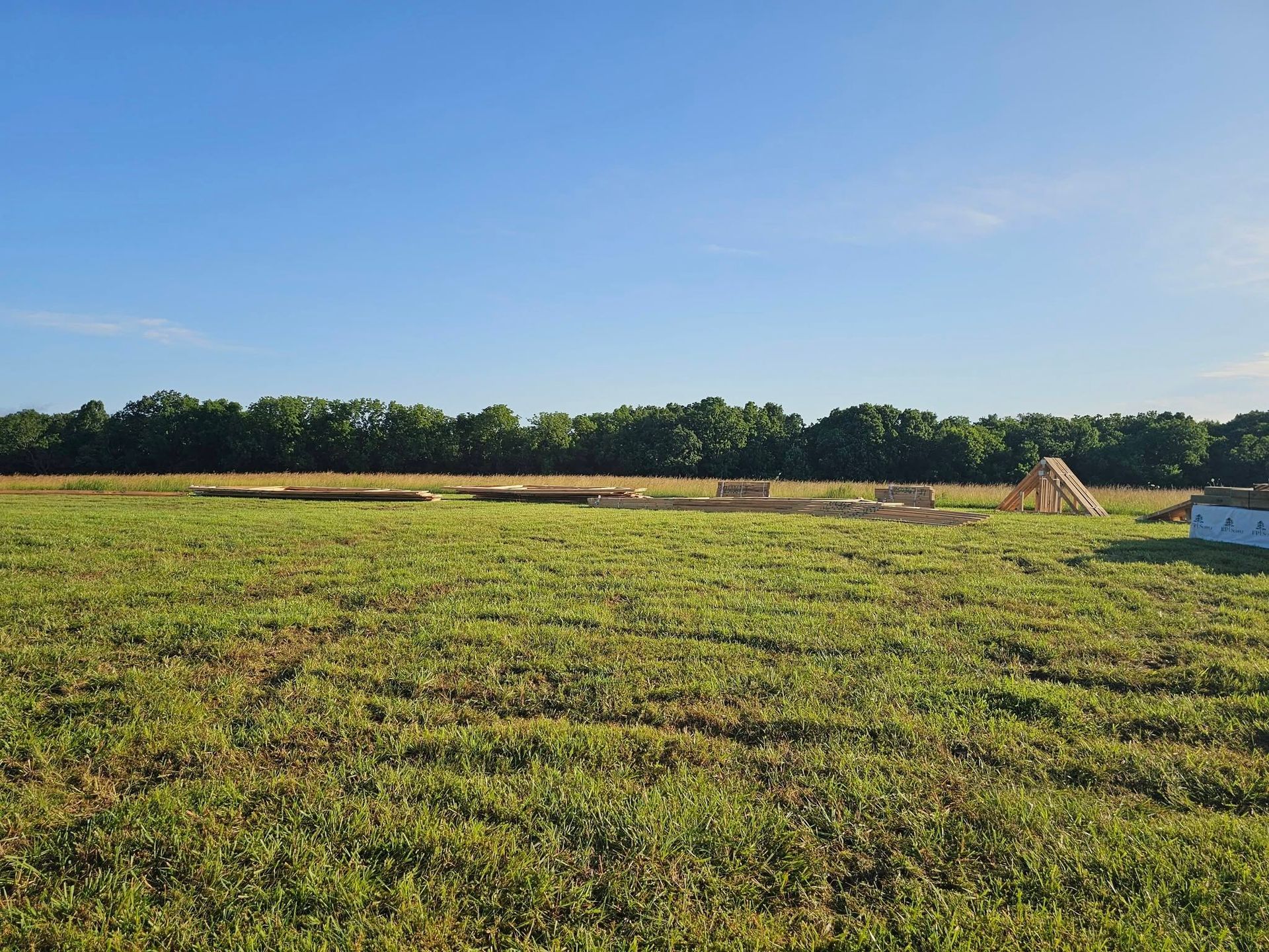 Wide grassy field under a blue sky with a tree line and small haystacks in the distance