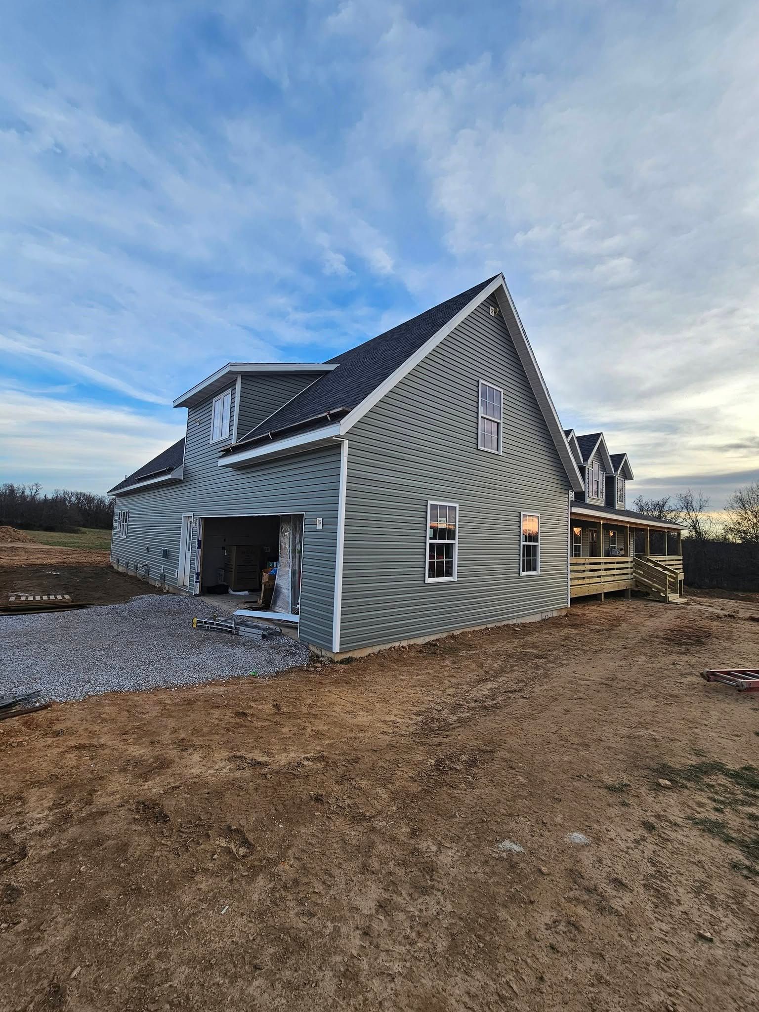 Gray house under construction with roof framing, gravel yard, and dirt foreground under a cloudy sky