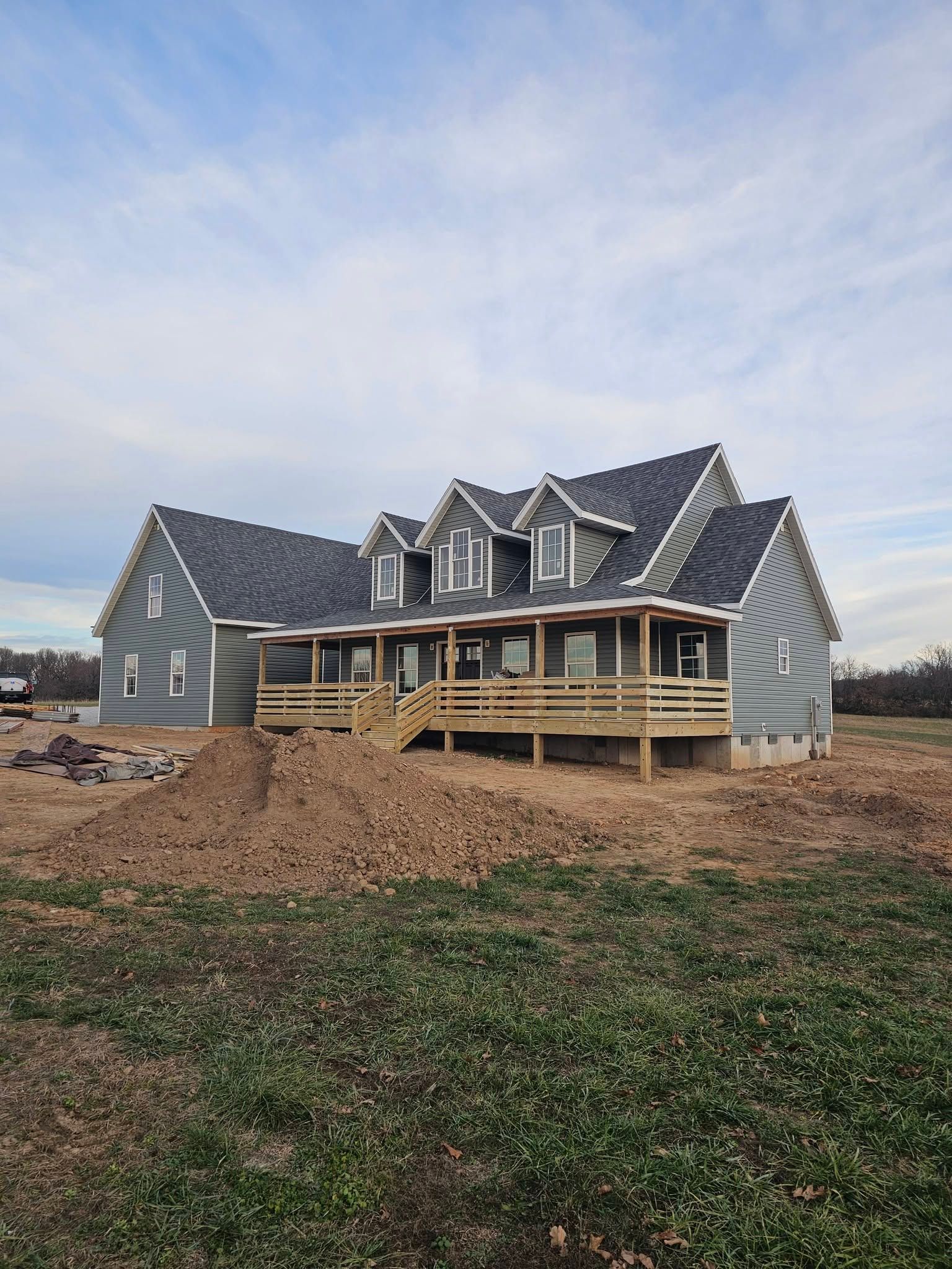 New house under construction with gray siding, dormer windows, and a wraparound porch on a dirt lot