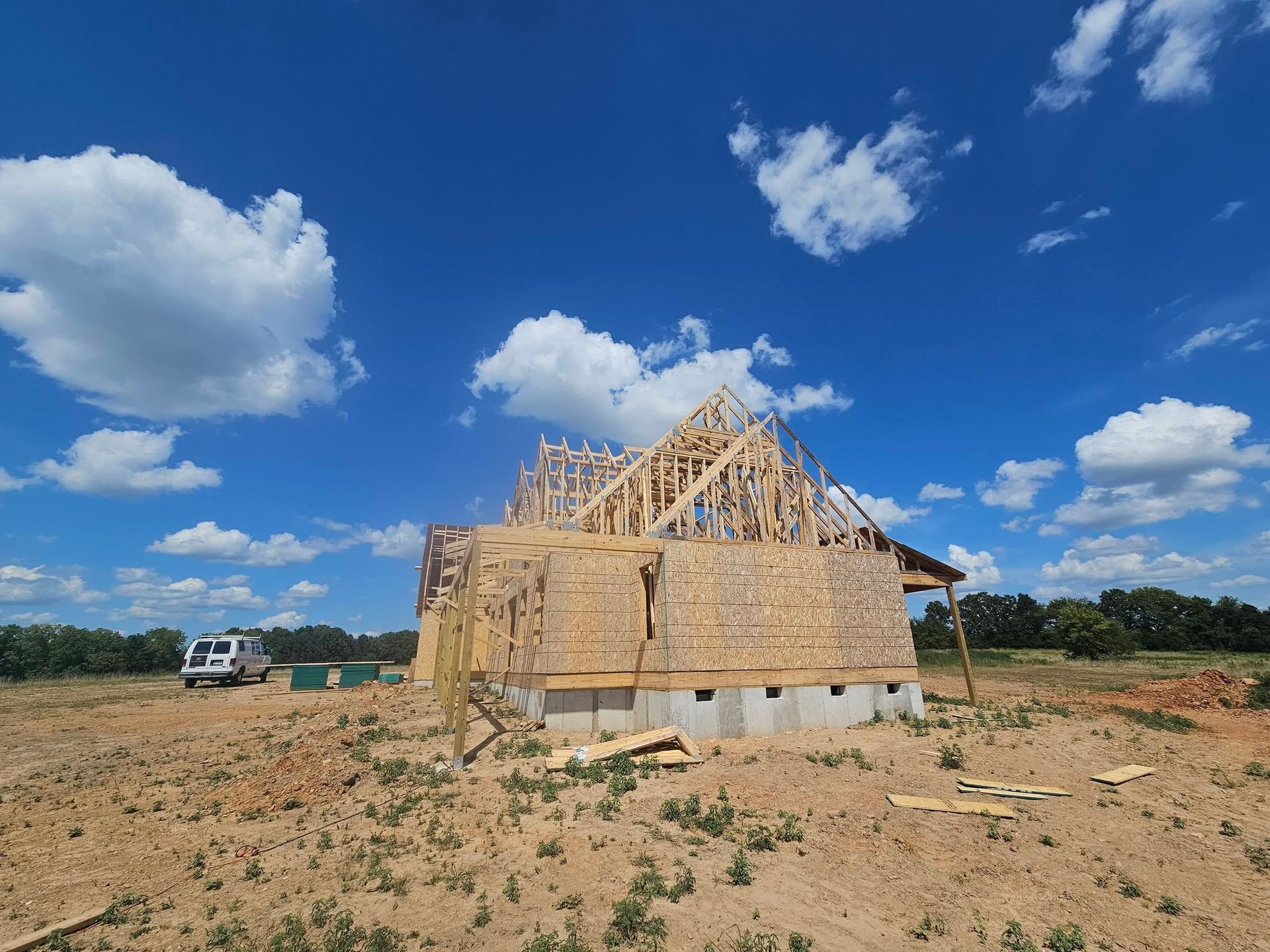 Partially built wooden house frame on a sandy lot under a bright blue sky with clouds