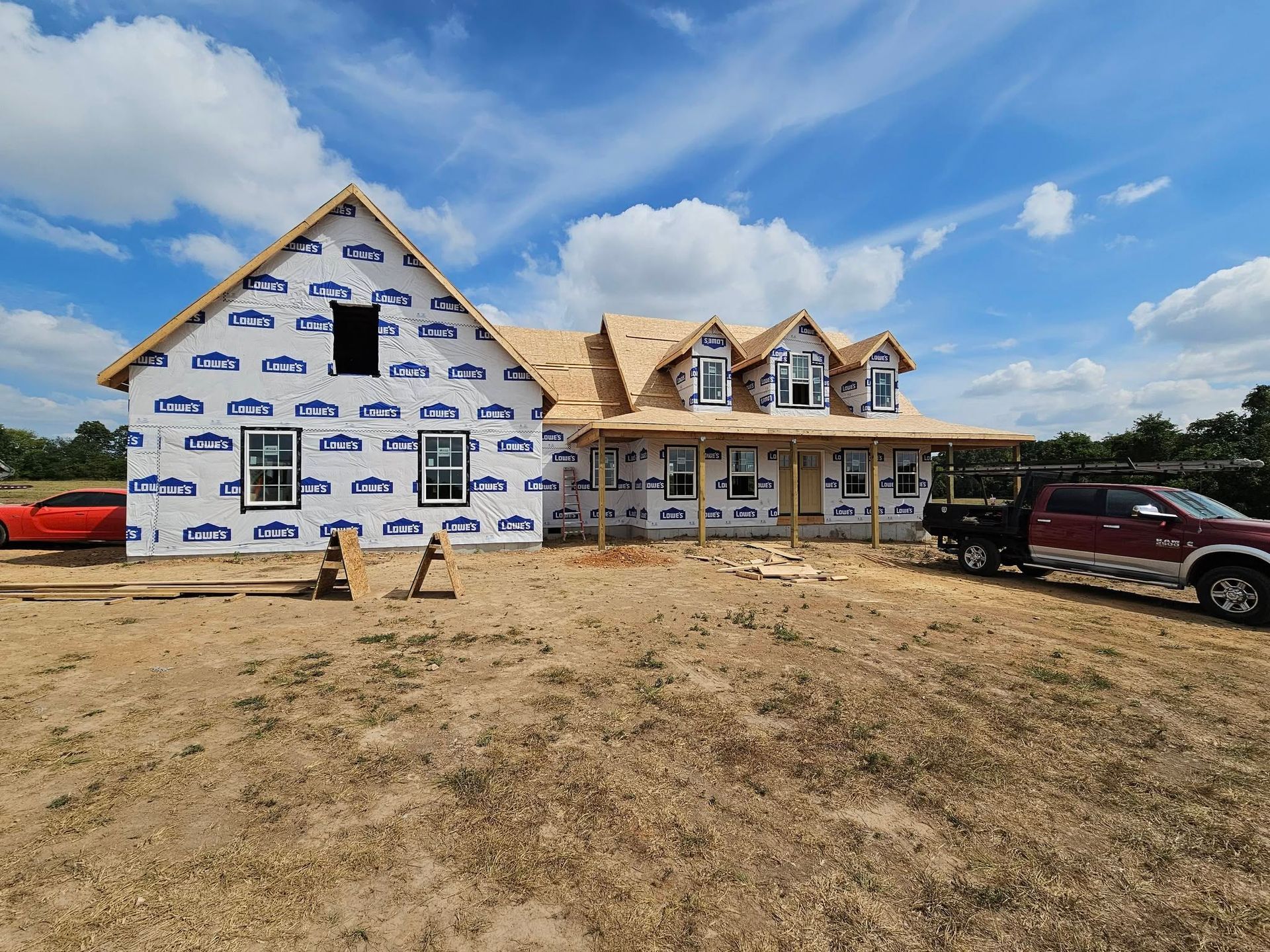 Partially finished house wrapped in blue housewrap under a cloudy sky, with trucks parked nearby.