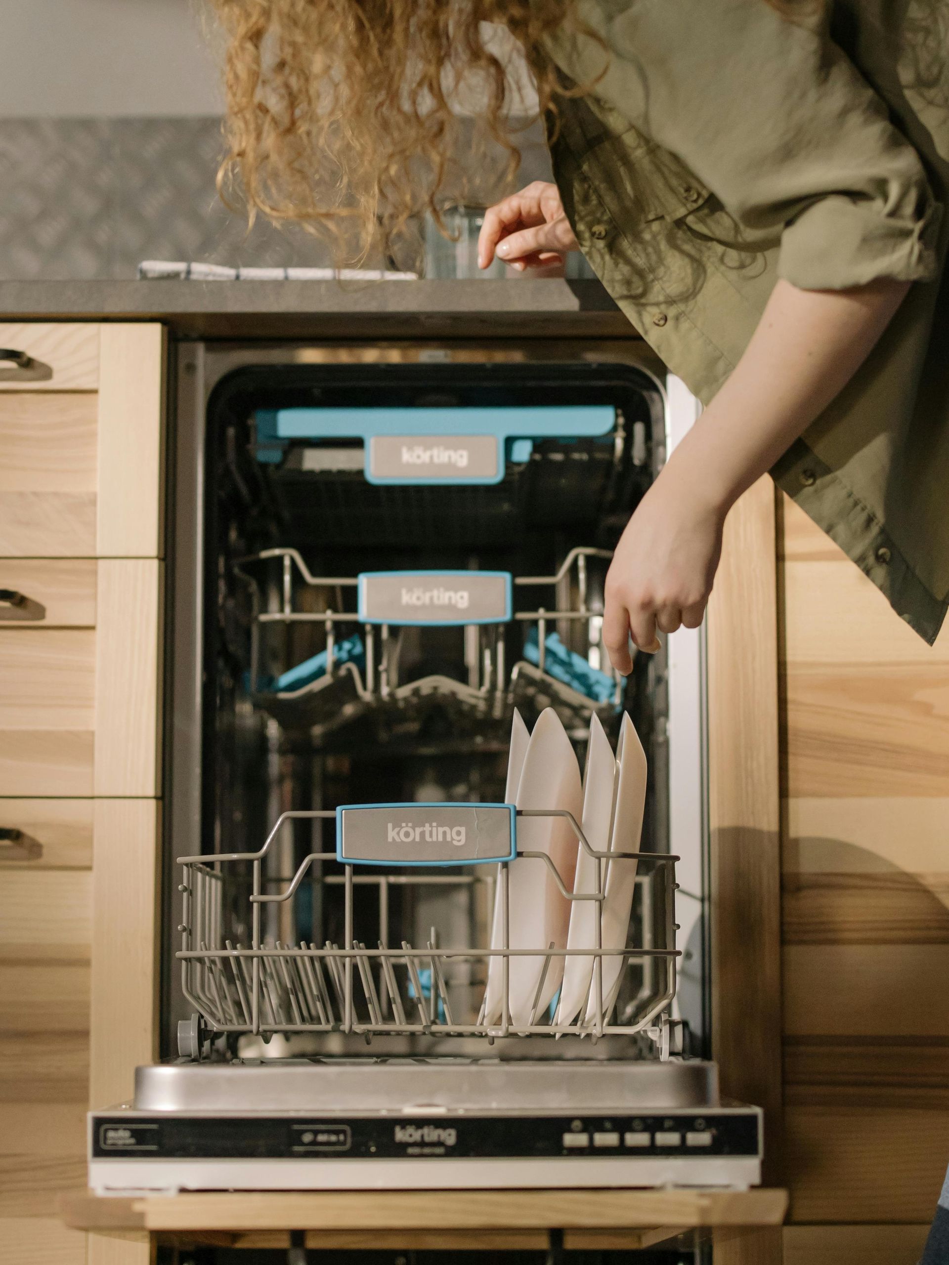Woman loading silverware into a dishwasher, set in a light wood kitchen.