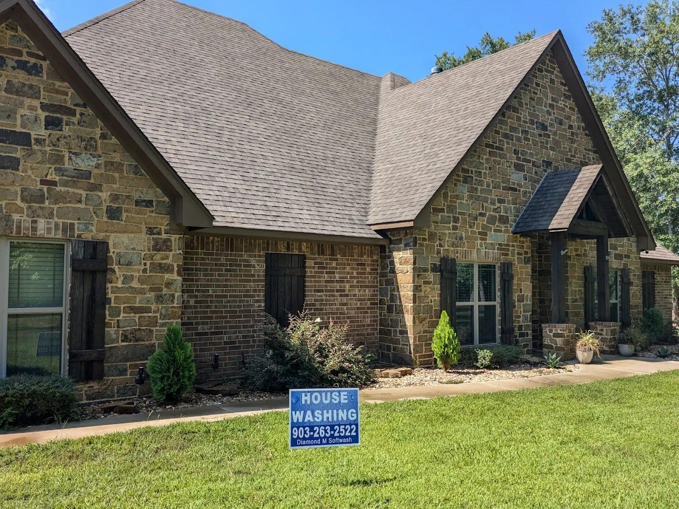 Stone house with shingled roof and front lawn, featuring a blue “House Washing” sign near the entrance.