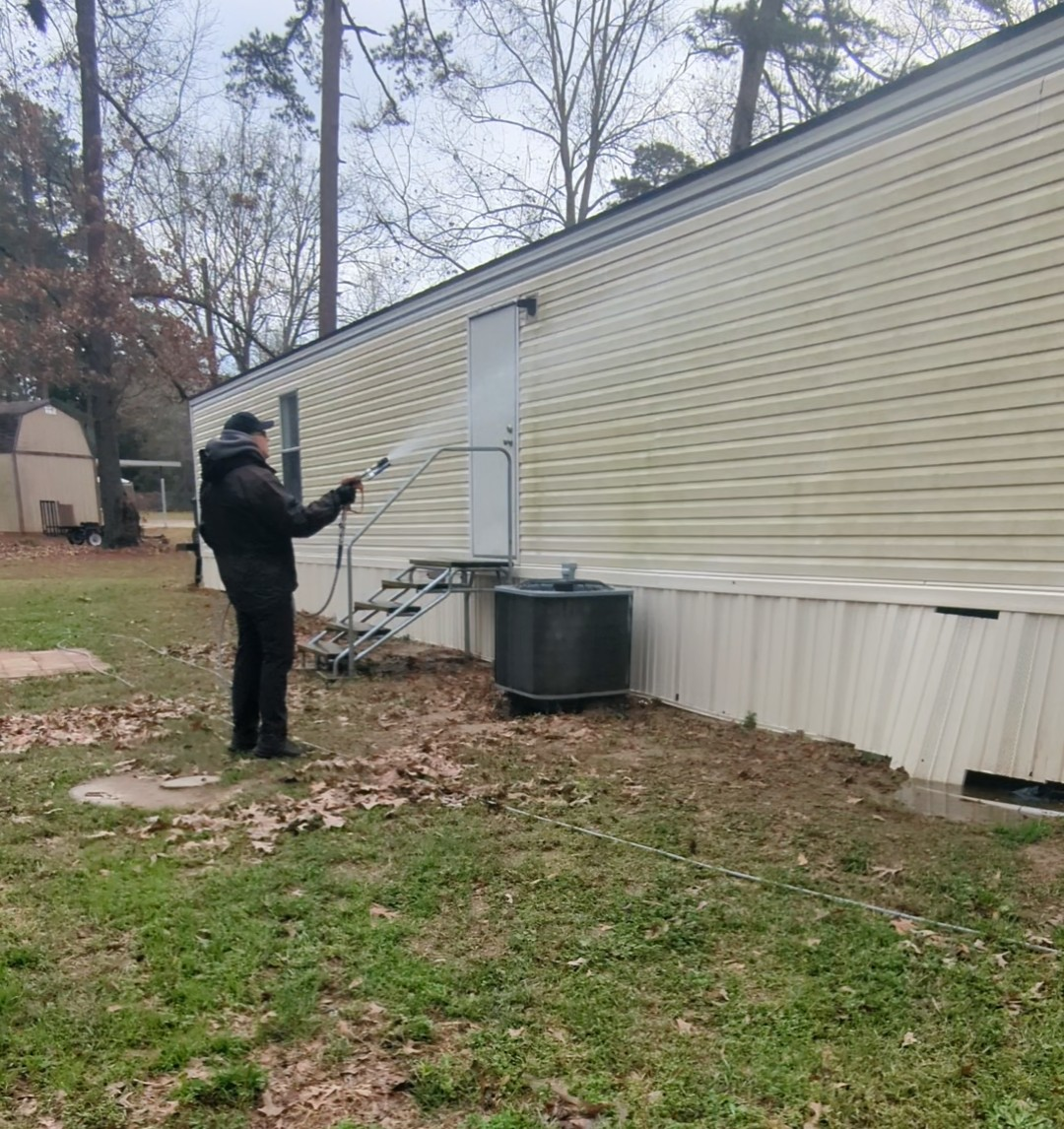 Person pressure-washing the side of a beige mobile home beside a small porch and yard