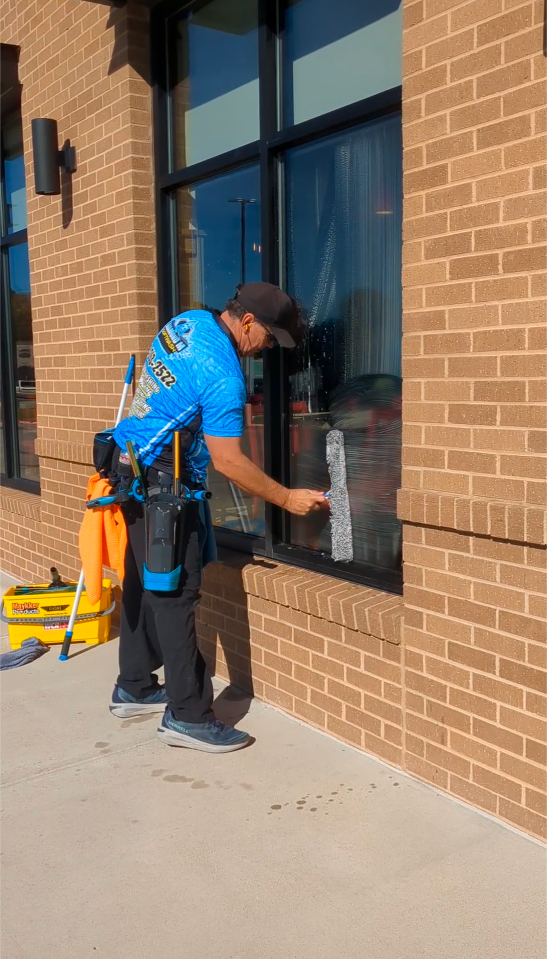 Window cleaner wiping a storefront window beside a brick wall, with cleaning tools on a cart.