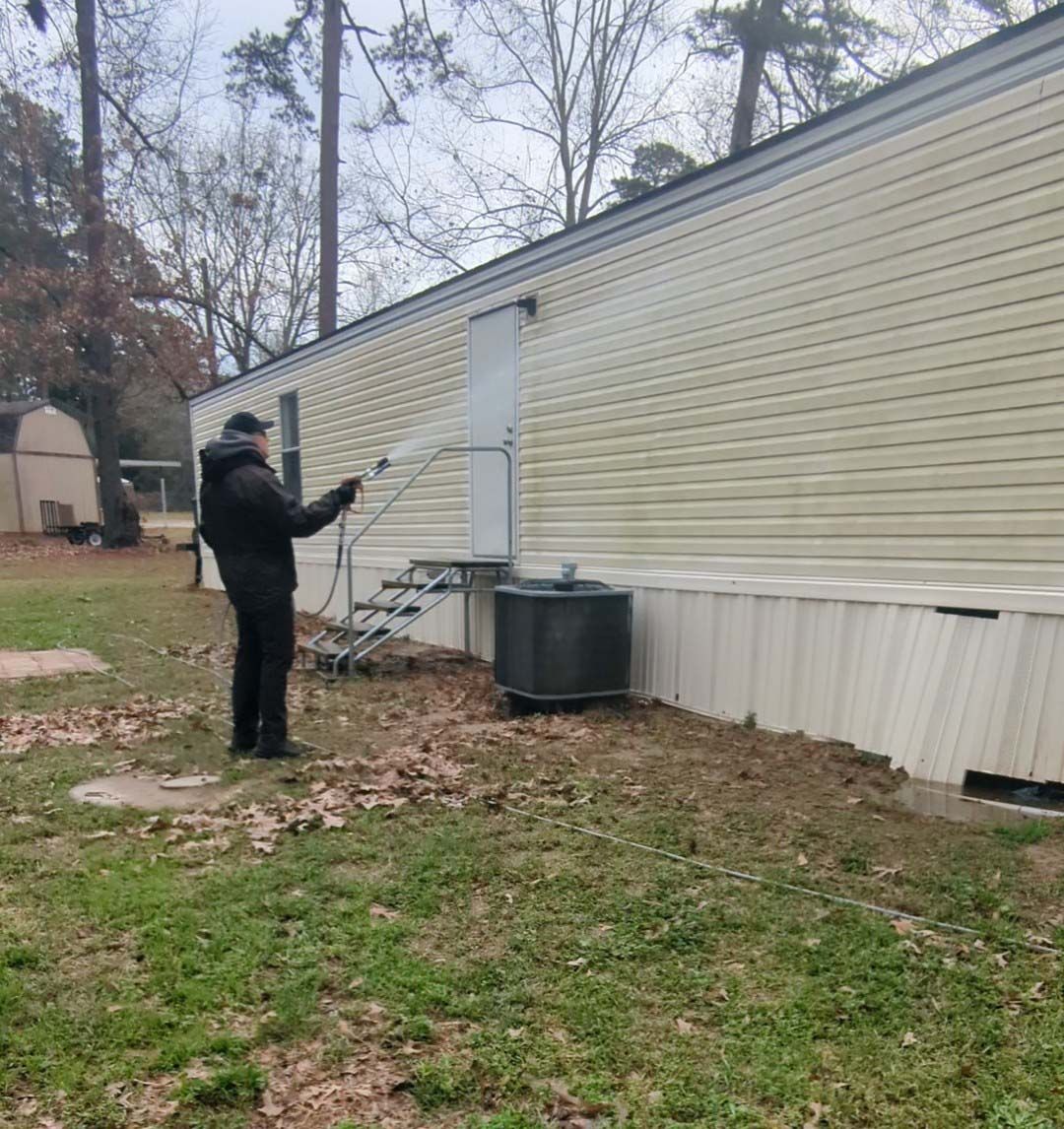 Person using a pole near a beige mobile home, standing in a grassy yard with a ladder and utility tank.