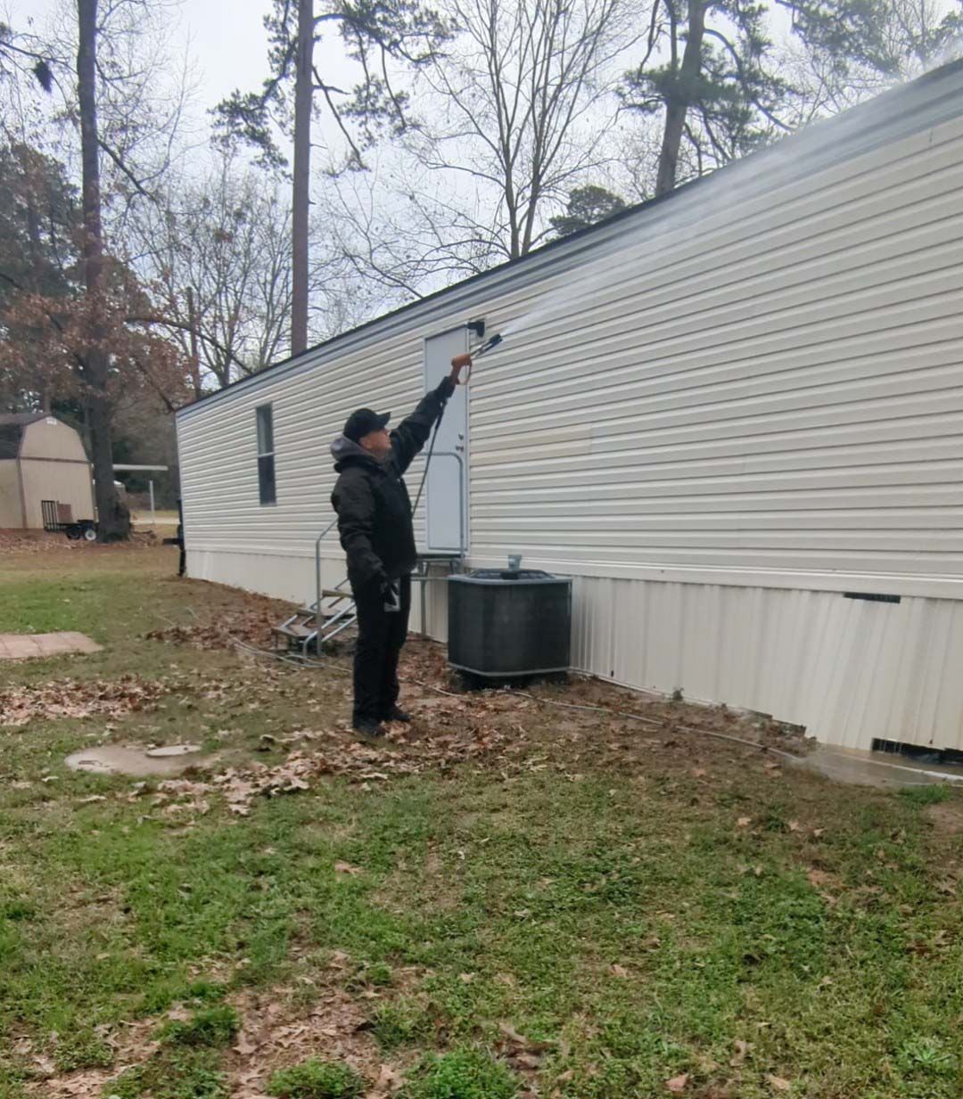 Person painting the side of a white mobile home with a long roller outdoors in a yard.