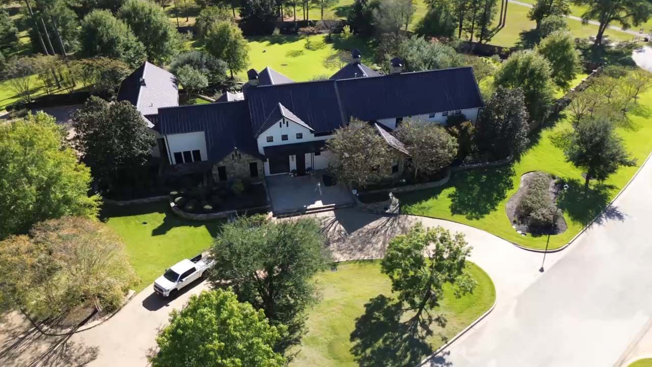 Aerial view of a large dark-roofed house with trees, lawn, and a driveway in a green neighborhood.