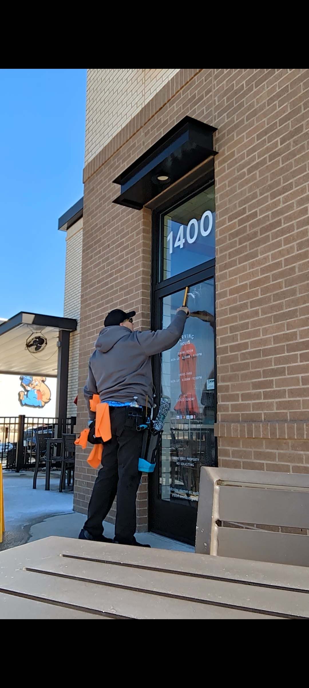 Worker on a ladder cleaning a brick storefront window under a black awning.