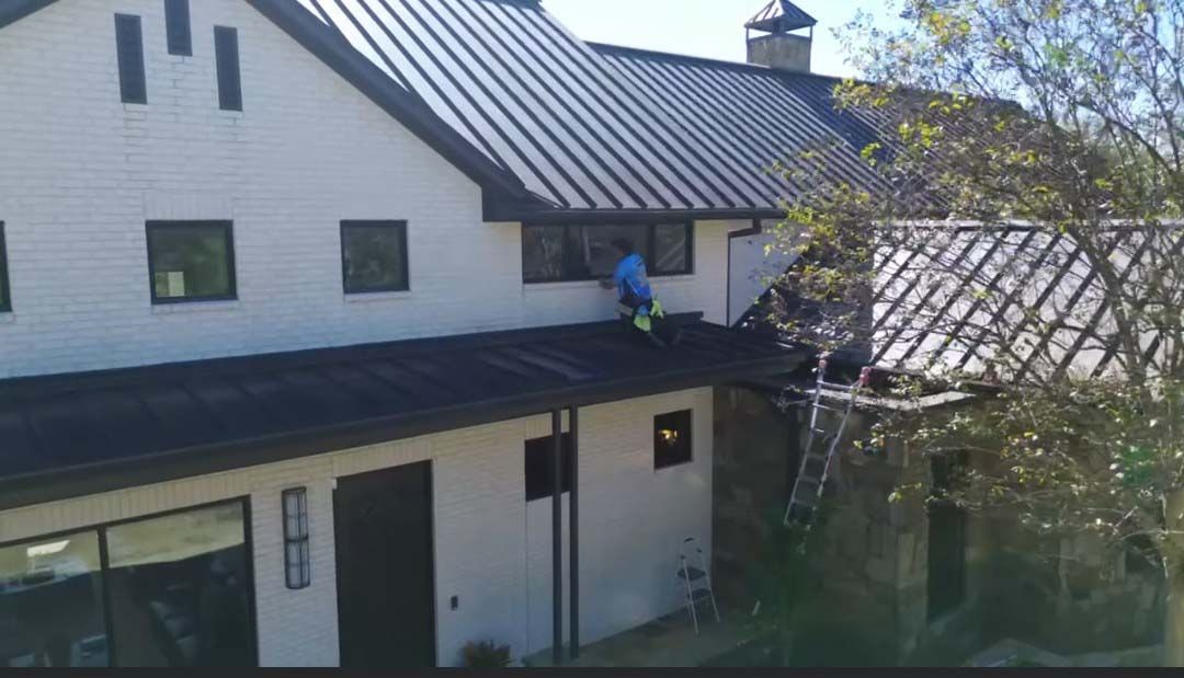 Person on a roof beside a white house with black trim, next to a ladder and tree branches