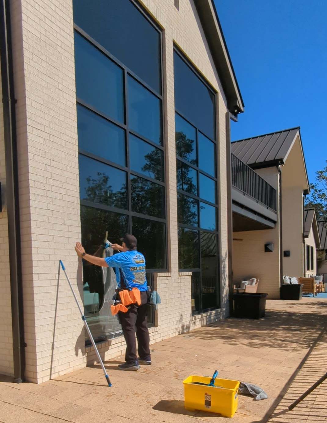 Worker cleaning tall windows on a brick building exterior, with a ladder and tools nearby.