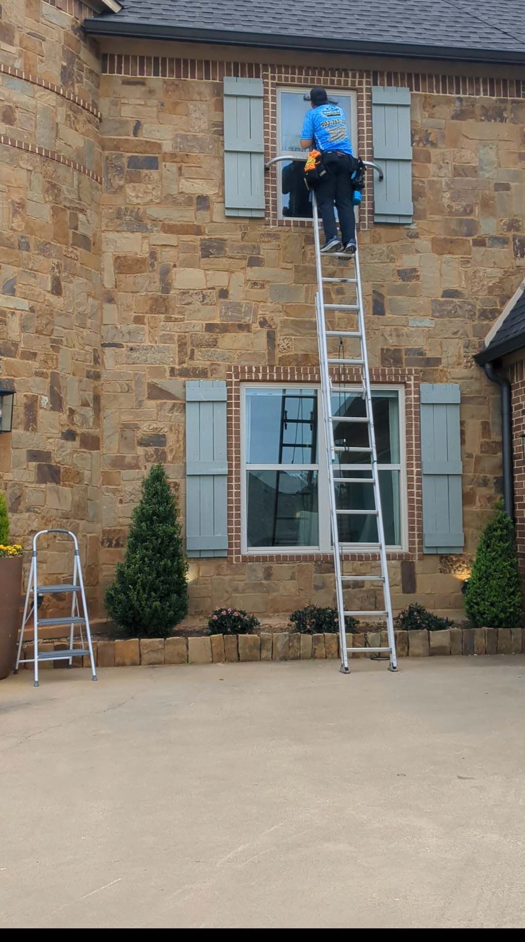 Worker on a ladder cleaning or repairing an upstairs window of a stone house