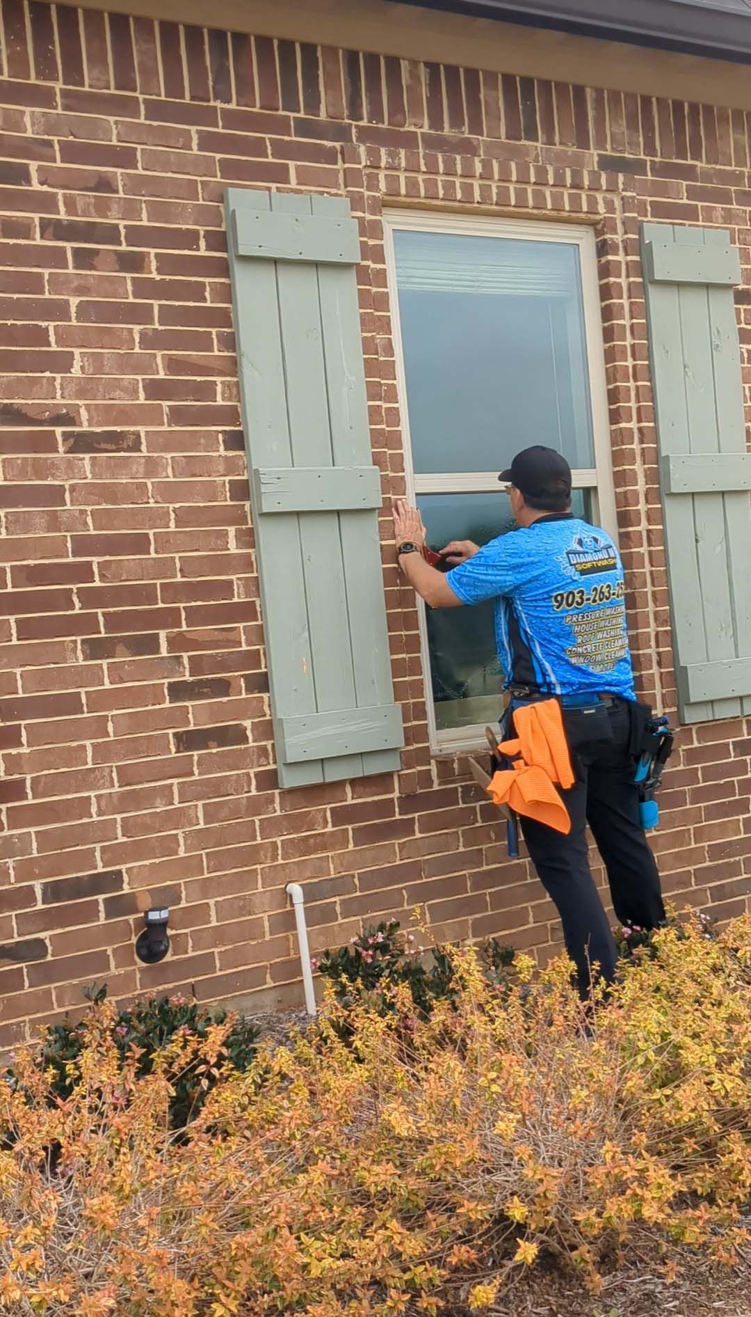 Person painting gray shutters on a brick house beside a window.