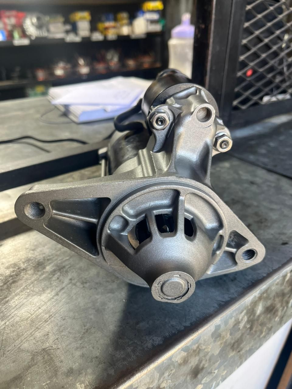 Primer plano de un motor de arranque metálico para automóvil sobre un banco de trabajo, con un garaje desenfocado al fondo.