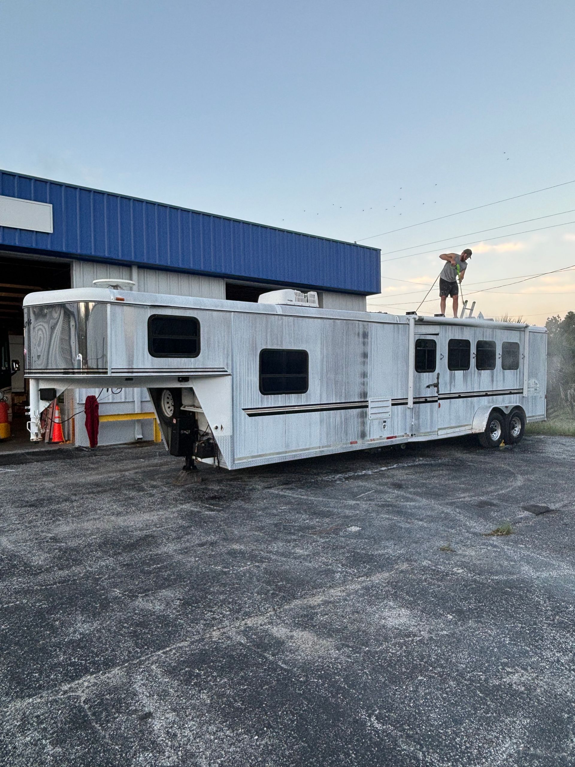 A horse trailer being cleaned by a person on the roof; parked in front of a blue building, gray pavement.