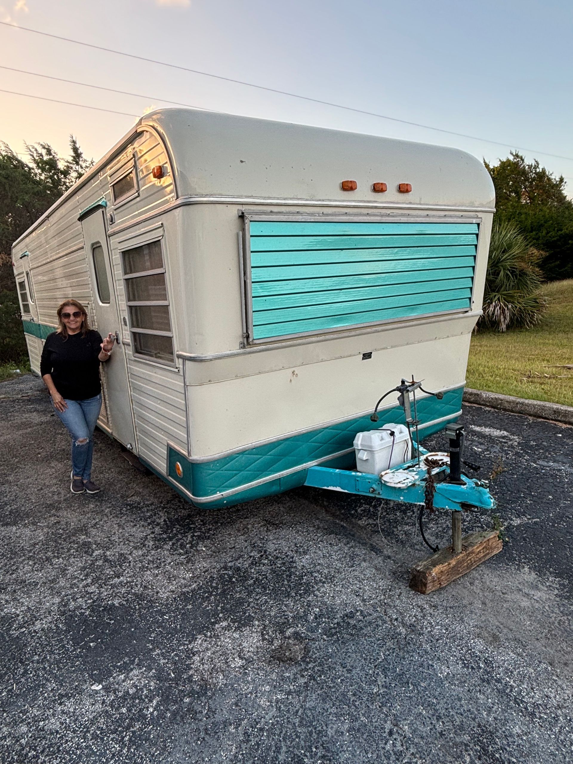 Woman stands next to a vintage teal and white travel trailer parked on asphalt.