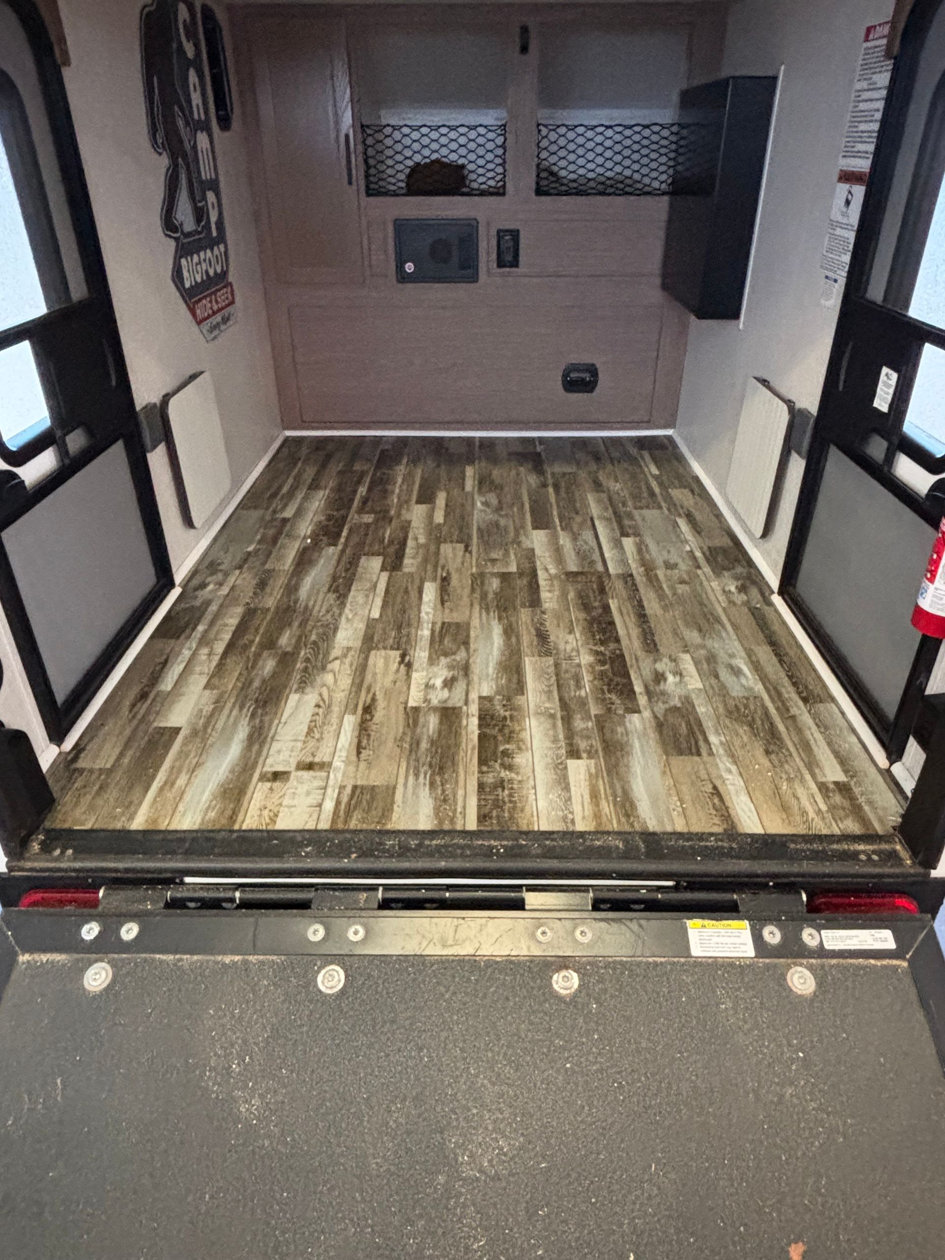 Inside of a cargo trailer with wood-look flooring. Ramp is down, doors open. Walls are neutral, items mounted.