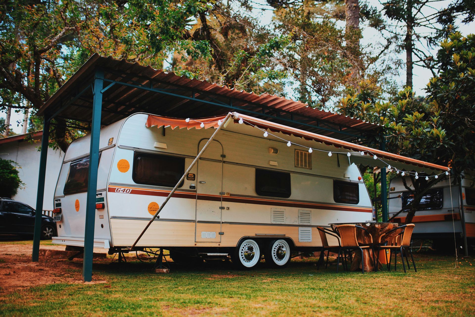 RV with awning, under a canopy in a grassy area, string lights, surrounded by trees.