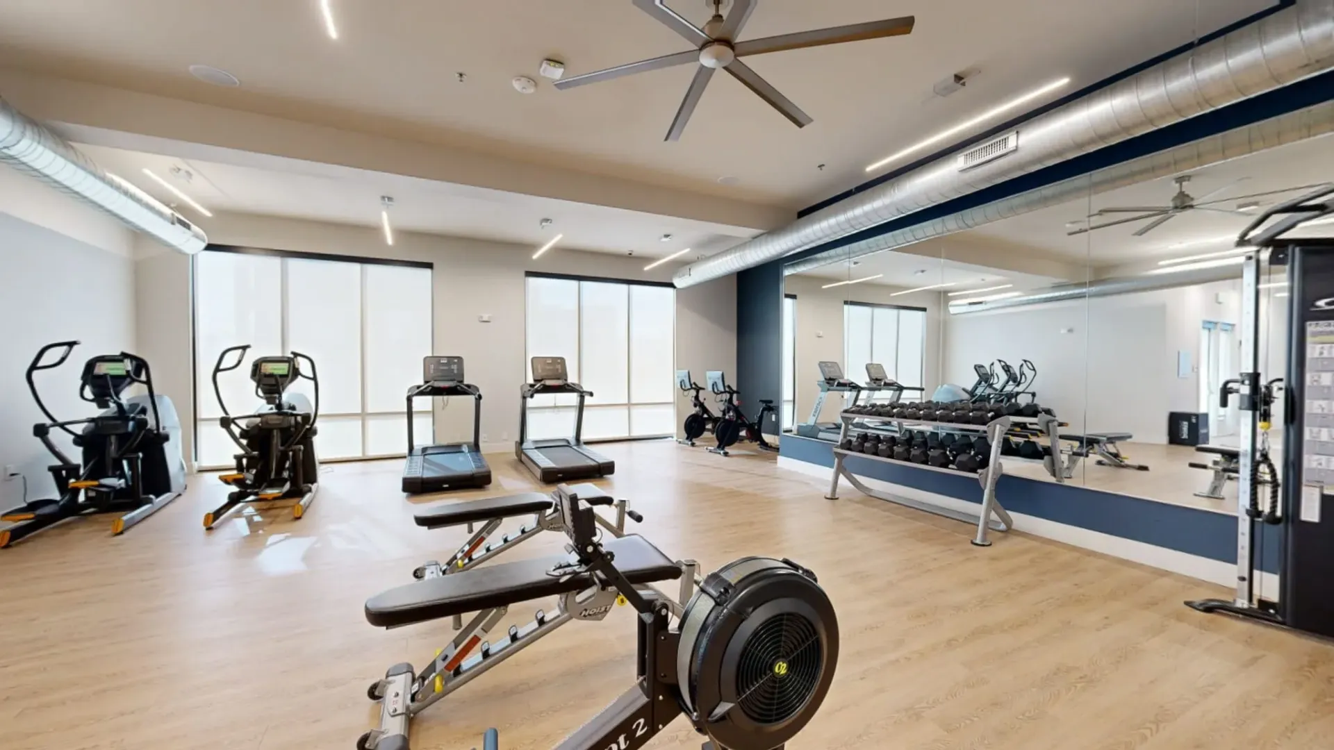 Well-lit community gym with cardio machines, free weights, and mirrors.