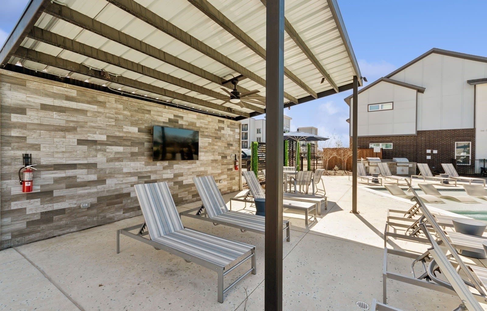 Outdoor pool deck with striped lounge chairs under a metal canopy and a wall-mounted TV.