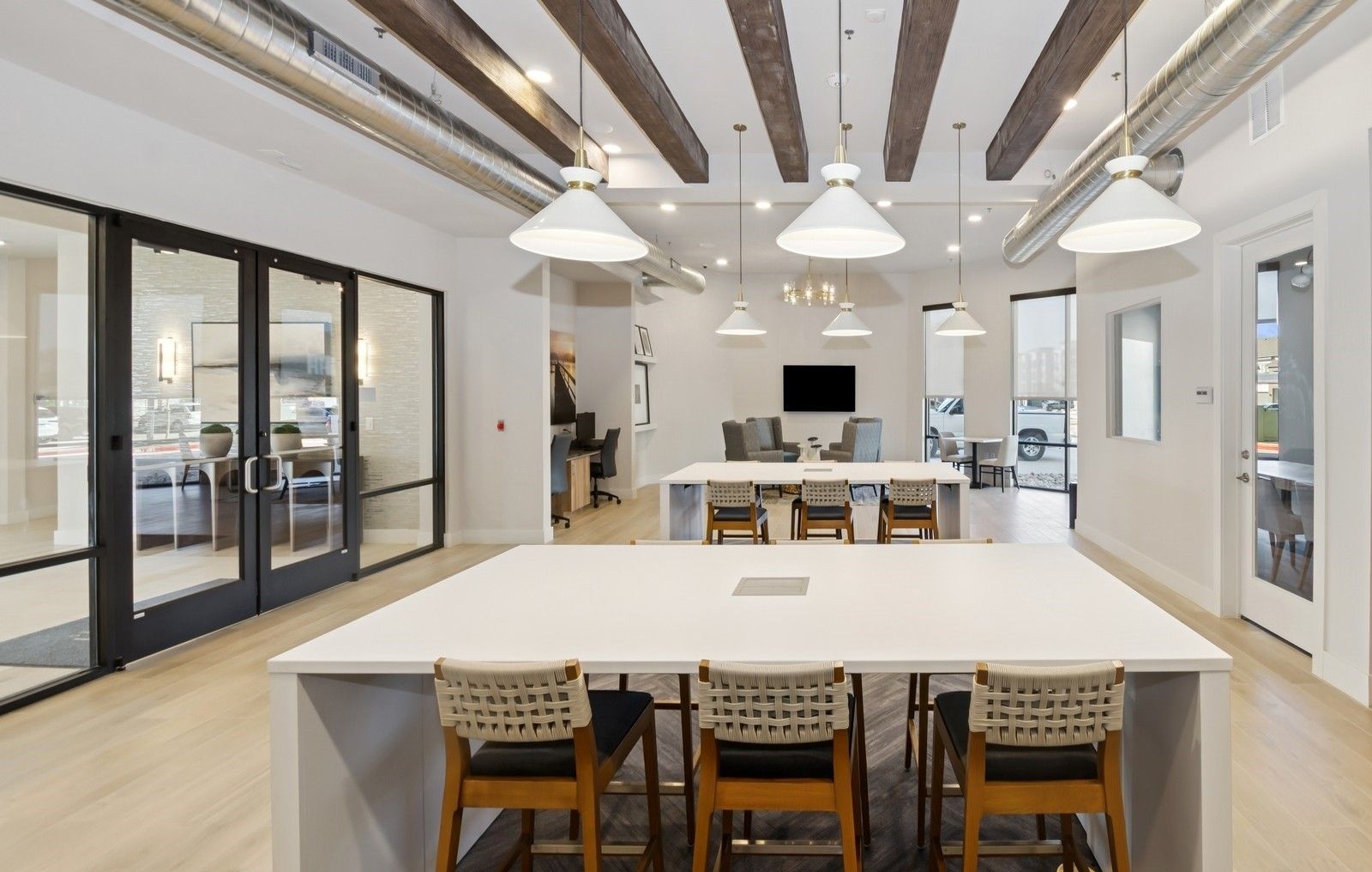 Interior communal lounge with long white tables, pendant lights, glass doors, and exposed ductwork.