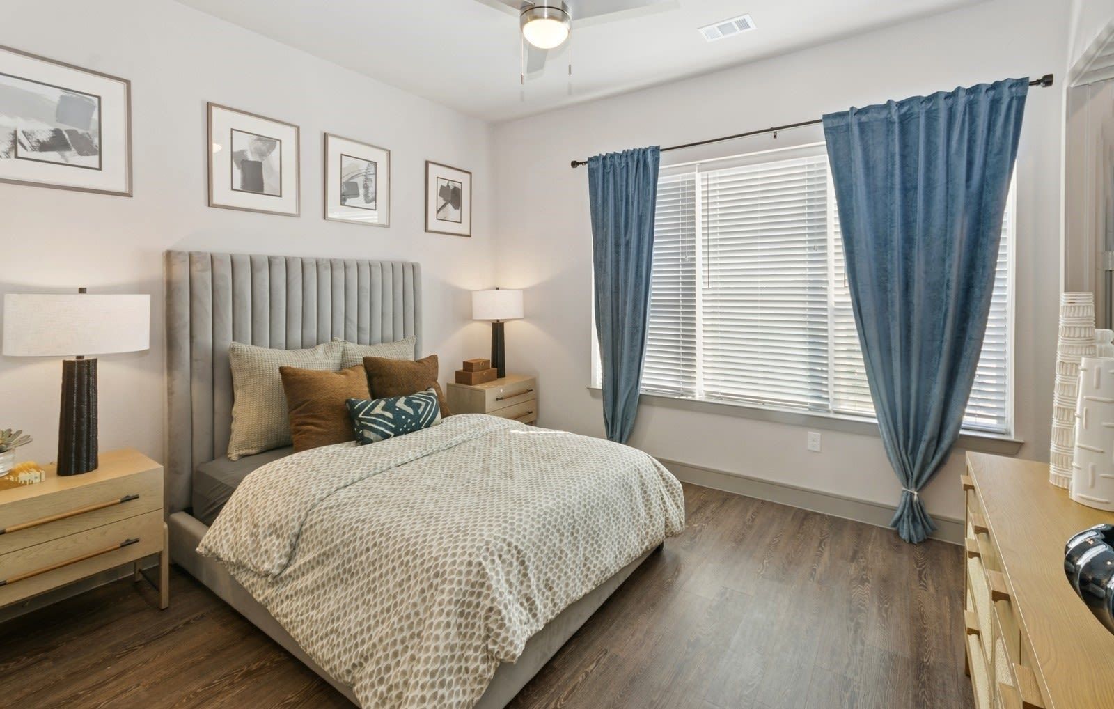Bedroom in apartment with upholstered headboard, two nightstands, dresser, and blue curtains by a window.