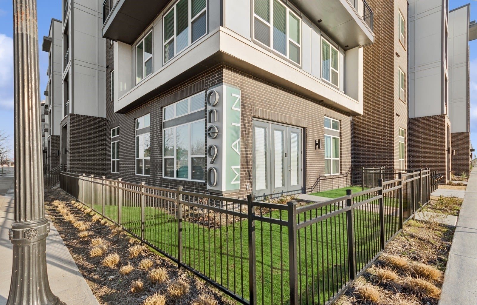 Exterior view of a modern apartment building with brick lower level, large windows, and a black metal fence.