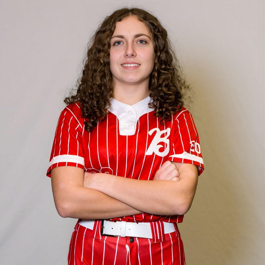 A person with curly hair wearing a red and white pinstriped softball uniform, standing with arms crossed against a backdrop.