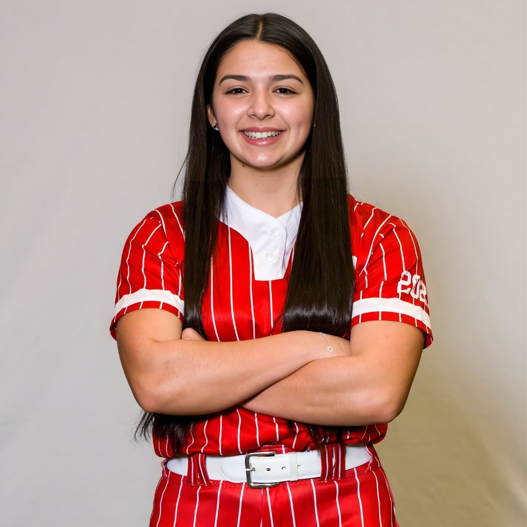 A smiling softball player with long dark hair wearing a red and white pinstriped jersey stands with arms crossed.