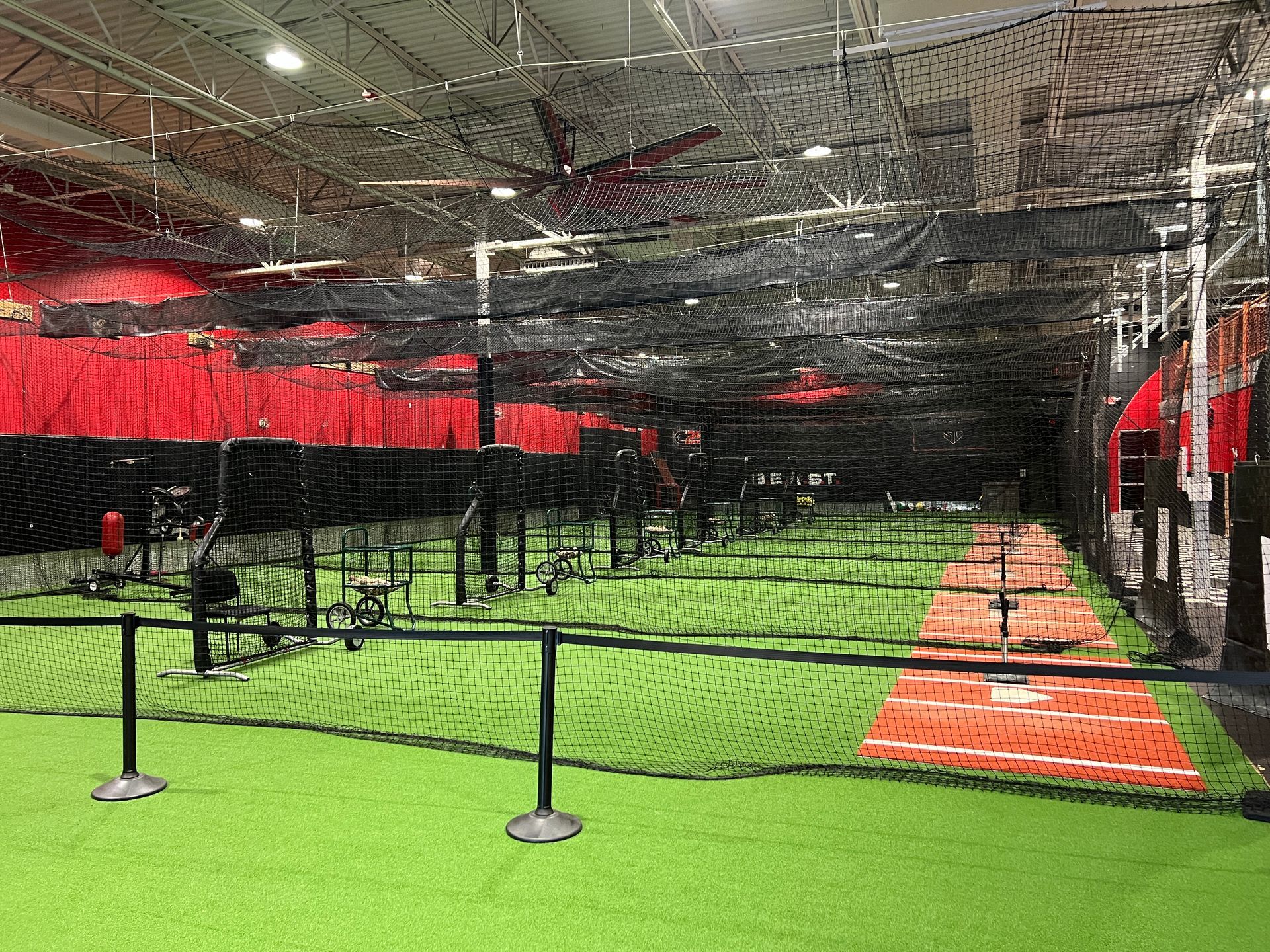 Indoor baseball training facility with multiple batting cages and netting over green turf and red dirt base paths.