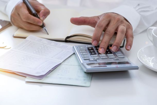 Person using a calculator, taking notes in a journal, and working with papers on a white surface.