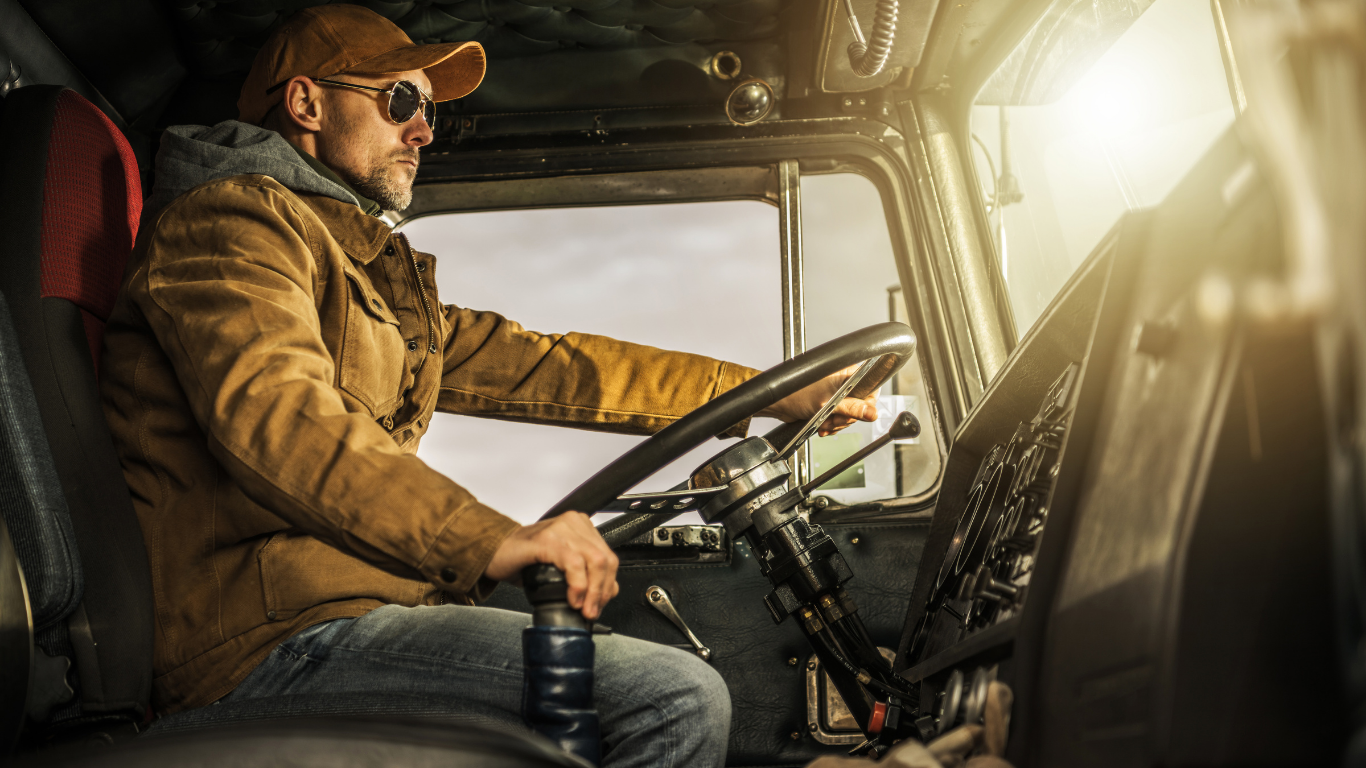 Truck driver at the wheel, wearing a cap and sunglasses, sunlight streams through the windshield.