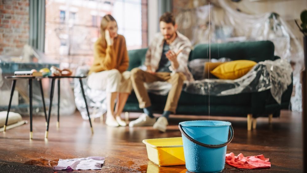 A man and a woman are sitting on a couch in a living room with a bucket of water on the floor.