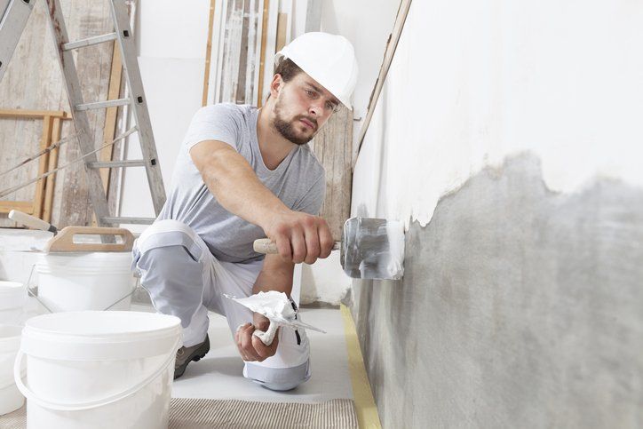 Man taking plaster from bucket and putting it on trowel to plaster the wall, wears helmet inside the building site of a house