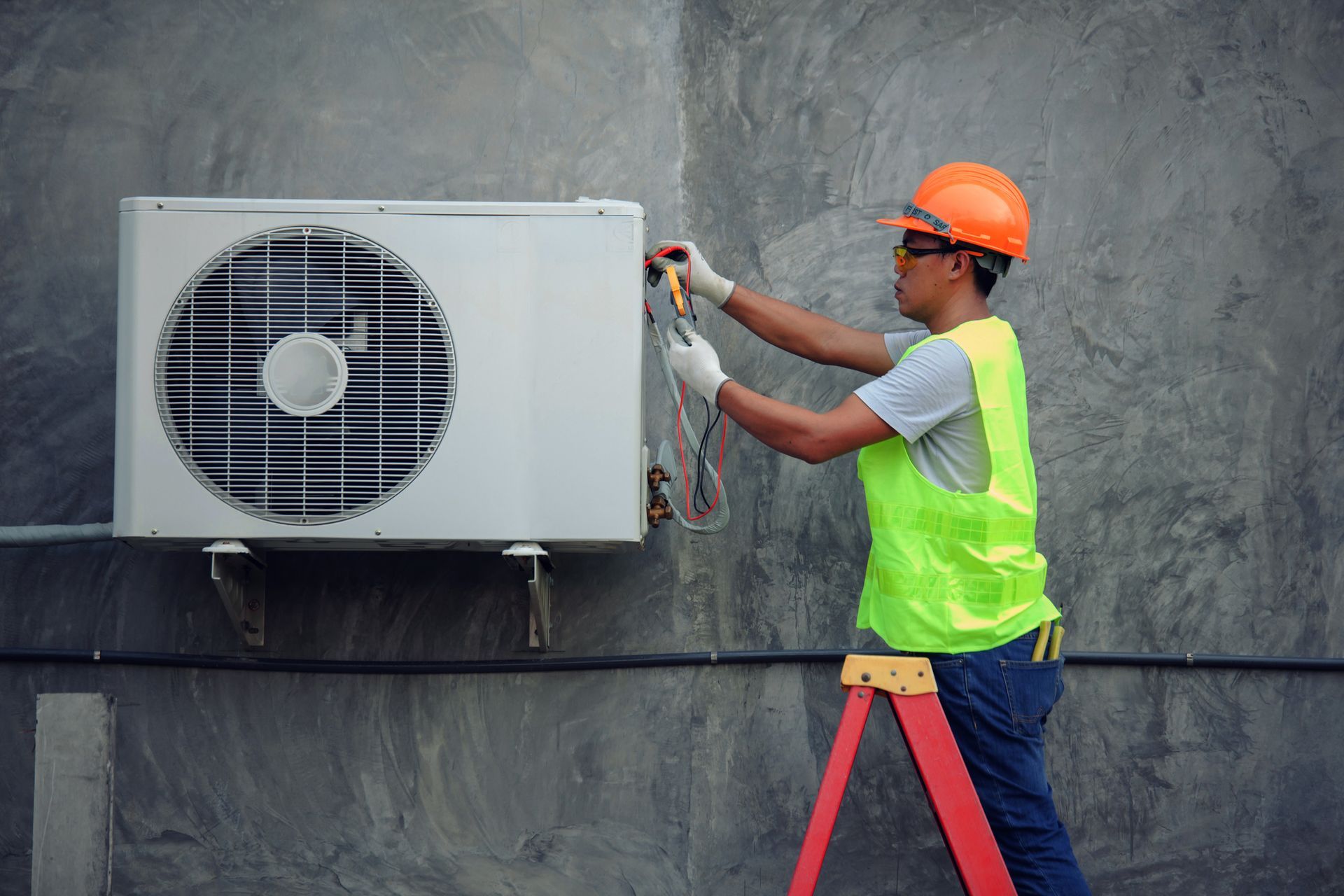 HVAC technician in hard hat on ladder, working on an AC unit attached to a grey wall.