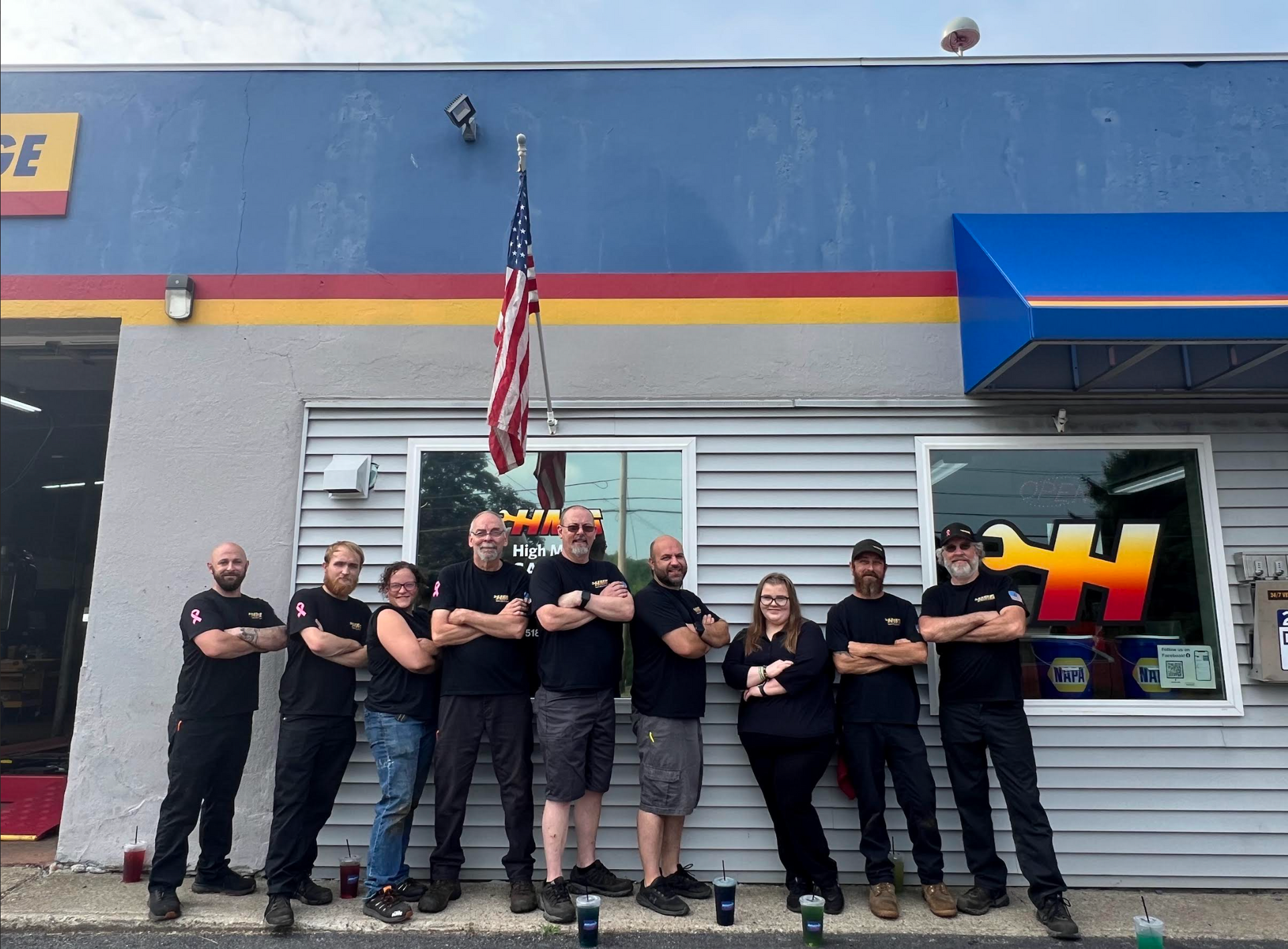 Group of people with arms crossed pose in front of a blue garage with an American flag.