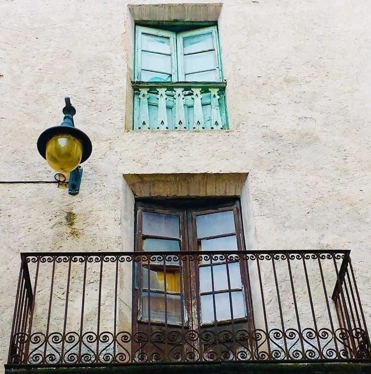 Dos ventanas con contraventanas color verde azulado y un balcón de hierro negro en un edificio de estuco color crema.