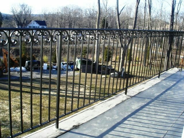 Una valla de hierro forjado negro proyecta sombras a lo largo de un sendero de hormigón junto a un jardín en invierno.