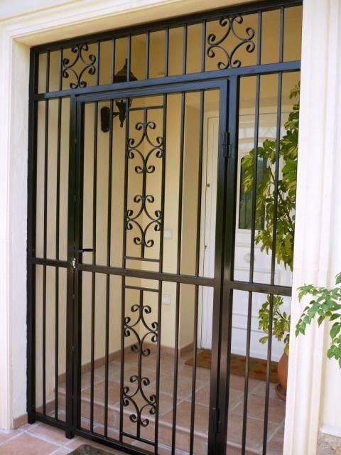 Puerta de seguridad de metal negro con detalles decorativos en forma de volutas en un marco de color beige.