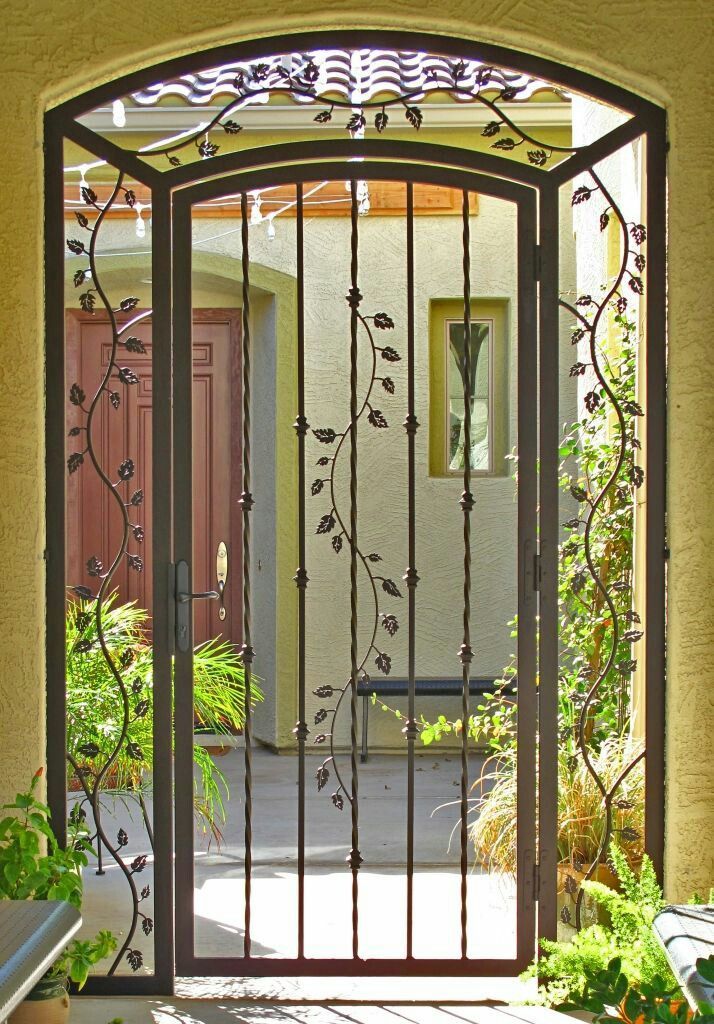 Una verja de hierro forjado enmarca la entrada a un patio soleado con plantas en macetas y una puerta principal al otro lado.