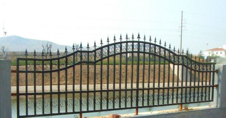 Una valla de hierro forjado negro con una puerta arqueada frente a una carretera y colinas bajo un cielo brumoso.