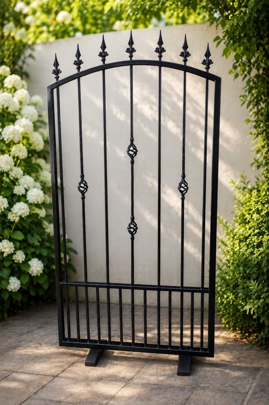 Puerta de jardín de hierro forjado negro con remates puntiagudos, situada entre flores blancas y setos verdes.