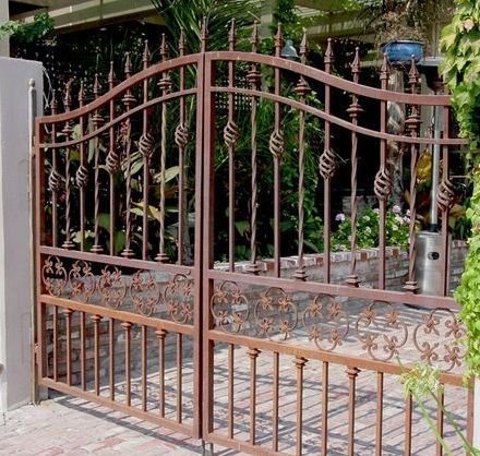 Puerta de hierro forjado color óxido con remate arqueado, situada frente a un jardín y un sendero de piedra.