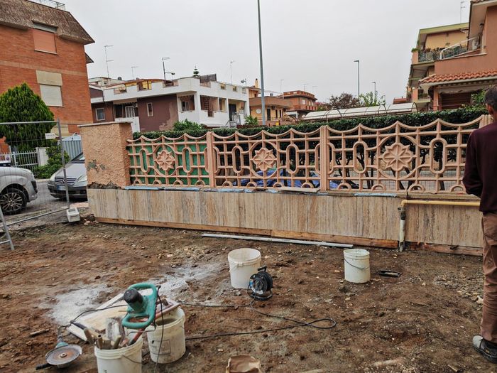 Pannello di recinzione in ferro battuto in un cantiere fangoso, con secchi e case sullo sfondo.