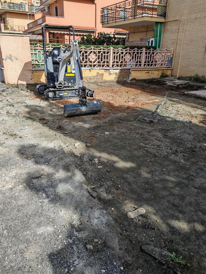 Escavatore che livella il terreno in un'area di costruzione accanto a un edificio arancione e a una recinzione.