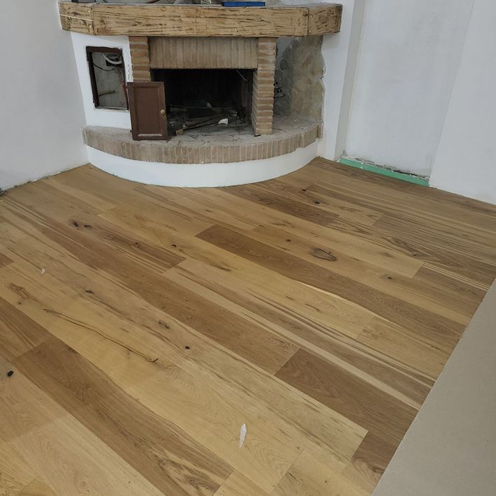 Stanza vuota con pavimento in legno chiaro e camino angolare in pietra contro pareti bianche.