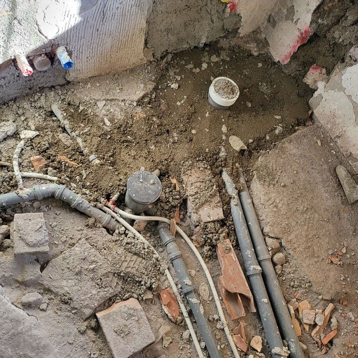 Scavo di costruzione con terra a vista, tubi e detriti accanto a un muro di cemento grezzo.
