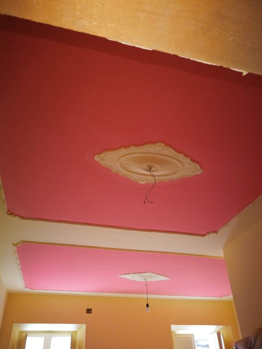 Soffitto con pannelli dipinti di rosa e rosso, medaglioni decorativi e un lampadario sospeso sopra una stanza.
