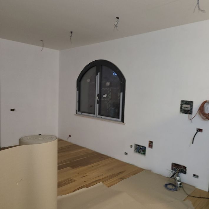 Stanza parzialmente rifinita con pareti bianche, finestra ad arco, cavi elettrici a vista e pavimento in legno grezzo.